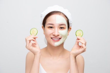 Beautiful young Asian woman with clay mask face holding cucumber piece over white background.の写真素材