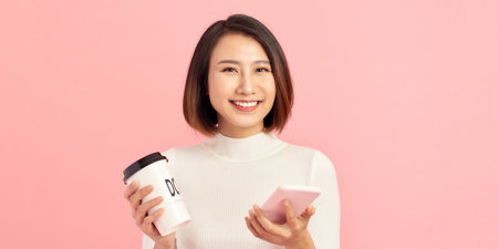 Young asian woman over isolated pink background holding coffee to take away and a mobileの写真素材