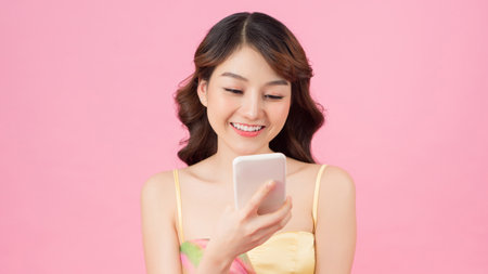 Portrait of a pretty smiling girl in summer clothes using mobile phone while standing isolated over pink backgroundの写真素材