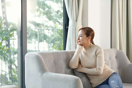Attractive peaceful young woman with cup of tea sitting and dreaming on sofa at home