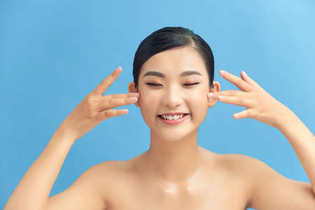 Beautiful smiling asian girl model with natural makeup touching glowing hydrated skin on blue background closeupの写真素材