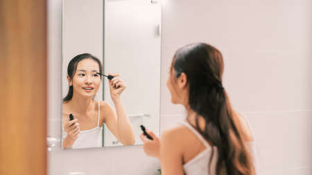 Pretty young woman putting some mascara on her eyelashes in front of a bathroom mirrorの写真素材