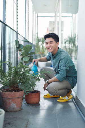 Attractive Young Man on Apartment Balcony Watering Plants in Box from Blue Watering Canの写真素材
