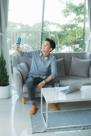 Selfie. Portrait of a young beautiful man wearing blue shirt in blue headphones takes a photo of himself on mobile phoneの写真素材