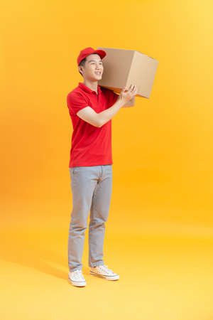 happy young delivery man in red cap standing with parcel post box isolated over yellow backgroundの写真素材