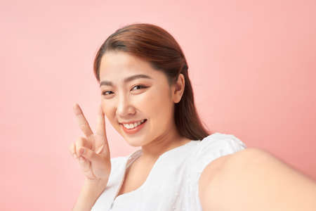 Young cheerful woman taking a self-portrait and gesturing v-sign against pink background