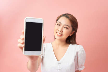 Young woman show display of mobile cell phone with black screen and smiling on a pink background. Focus on hand with mobile phoneの写真素材