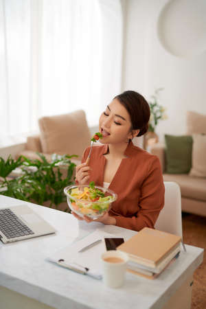 Portrait of woman watching movie on laptop and eating vegetable salad at table in breaktimeの写真素材