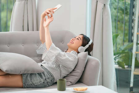 Happy young relaxed woman with headphones sitting on couch and listening to meditation music at leisureの写真素材