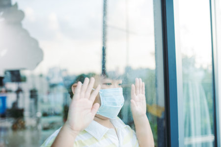 Cute asian young boy wearing medical mask, standing idle in front of close window, looking through...