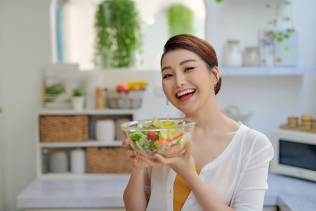 Smiling young woman eating fresh salad in modern kitchenの写真素材