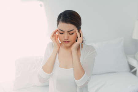 Miserable young woman touching temples sitting on bed at home,の写真素材
