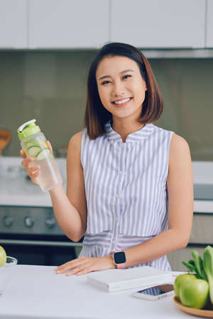 Beautiful young Asian woman holding detox drink in kitchen. Healthy drink, weight loss food concept.の写真素材