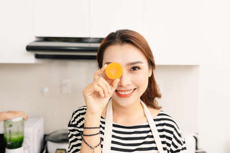 Young beautiful woman in orange apron sitting at table, having fun, carrot on eyeの写真素材