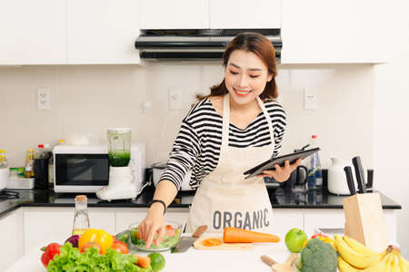 Closeup portrait of smiling young beautiful woman using tablet computer, reading recipe and cooking in kitchenの写真素材