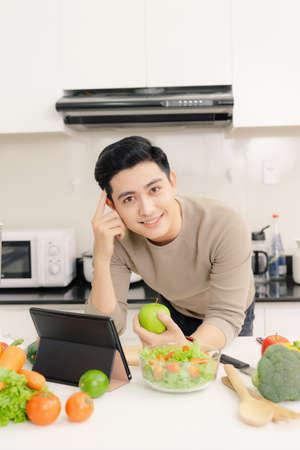 Young asian man holding apple fruit and using tablet while standing in kitchen roomの写真素材