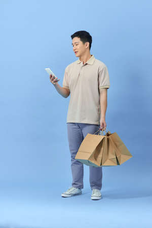 Young Asian man with shopping bags is using a mobile phone and smiling while doing shopping on blue backgroundの写真素材