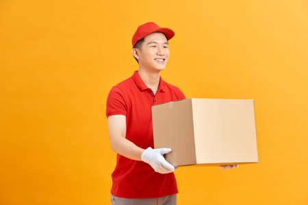 Image of a happy young delivery man in red cap standing with parcel post box isolated over yellow background.の写真素材
