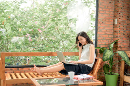 Attractive smiling young asian business woman relaxing on couch at home, working on laptop computer, talking on mobile phoneの写真素材