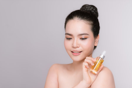Beauty portrait of an attractive young woman standing isolated over gray background, showing bottle with facial oilの写真素材