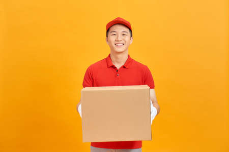 Image of a happy young delivery man in red cap standing with parcel post box isolated over yellow background.の写真素材