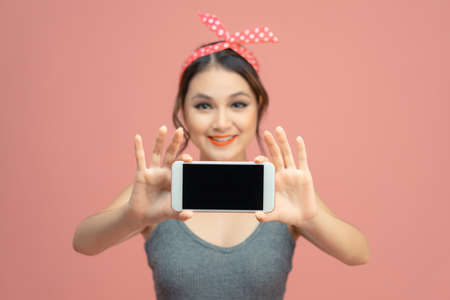 Smiling asian woman showing blank screen mobile phone while standing isolated over pink backgroundの写真素材