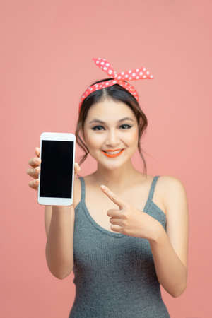 Portrait of a smiling casual girl pointing finger at blank screen mobile phone isolated over pink backgroundの写真素材
