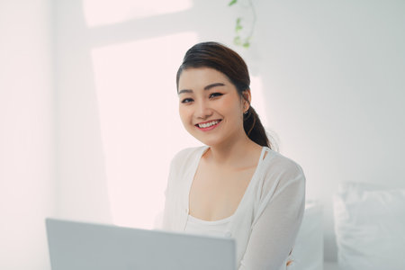 Happy casual beautiful woman working on a laptop sitting on the bed in the house.の写真素材