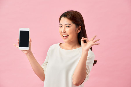 Happy Asian portrait beautiful cute young woman excited holding mobile phone and gesturing ok sign, studio shot isolated on pink backgroundの写真素材