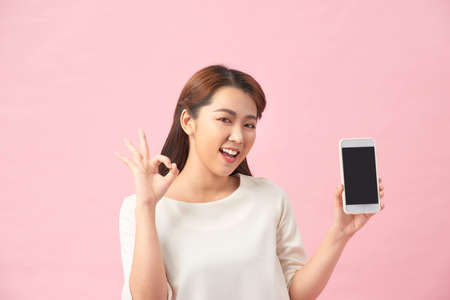 Happy Asian portrait beautiful cute young woman excited holding mobile phone and gesturing ok sign,...