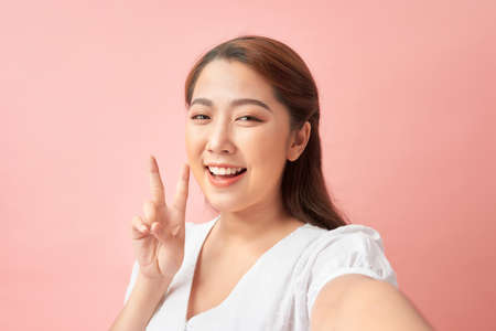 Young cheerful woman taking a self-portrait and gesturing v-sign against pink backgroundの写真素材