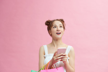 A portrait of young asian woman with colourful shopping bags enjoy mobile technology isolated on pink background.の写真素材