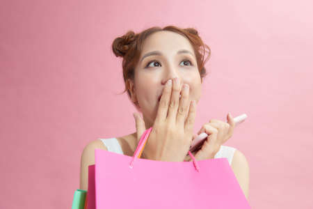 Excited asian shopaholic girl using phone posing with shopping bags on pink background. Sales and discounts season.の写真素材