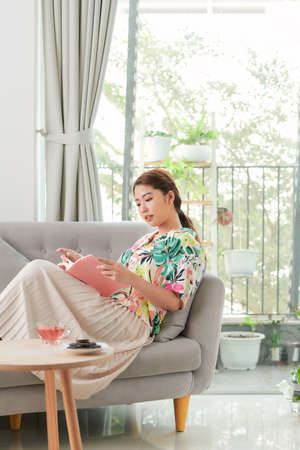 A young woman is reading a book while sitting on a sofa.の写真素材
