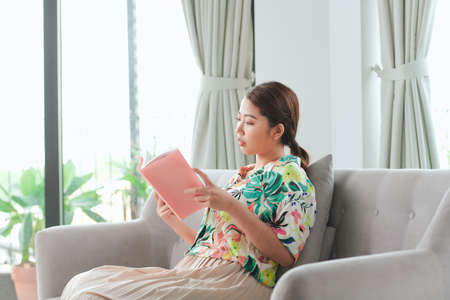 Casual young woman reading storybook on couch at homeの写真素材