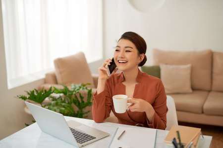 Young asian business woman holding mug and using smartphone while sitting in office desk with laptopの写真素材