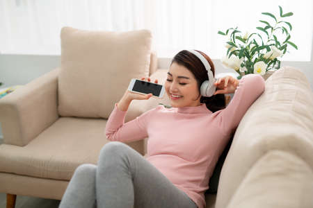 Happy young Asian woman in earphones singing and listening to music while sitting on sofa and holding mobile phone at home.の写真素材