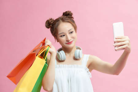 Asian beautiful happy young woman holding smartphone for selfie and shopping online with shopping bags on pink background.の写真素材
