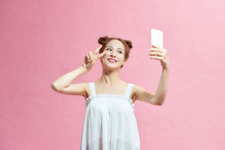Portrait of a young attractive woman making selfie photo on smartphone isolated on a pink backgroundの写真素材