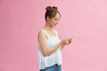 Happy attractive young woman standing and using mobile phone over pink backgroundの写真素材
