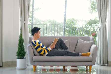 Young man reading book while sitting on the couchの写真素材