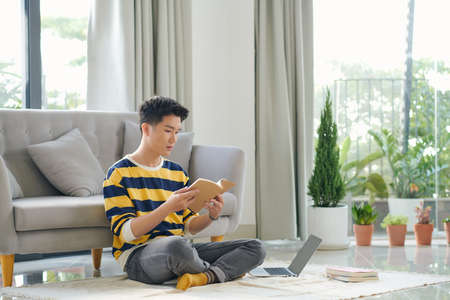 Asian man is sitting on the floor with a book and laptopの写真素材