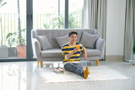Young male sitting on the floor in the living room and having video call via phoneの写真素材