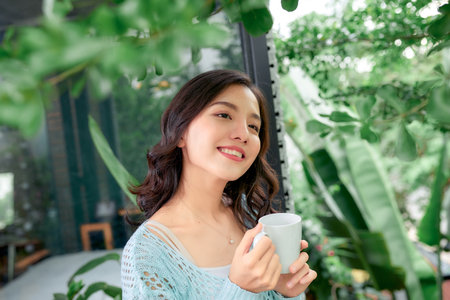 Beautiful young blonde woman in white dress drinking coffee, standing on the balcony on summer dayの写真素材
