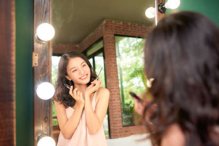 Cheerful attractive lady in homewear sitting in bedroom, using mascara, getting ready before going outの写真素材