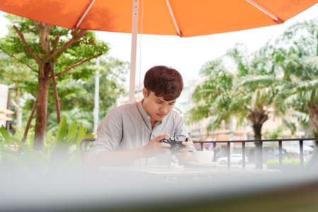 Successful hipster guy waiting friend in coffee shop while checking photos at vintage camera during free timeの写真素材