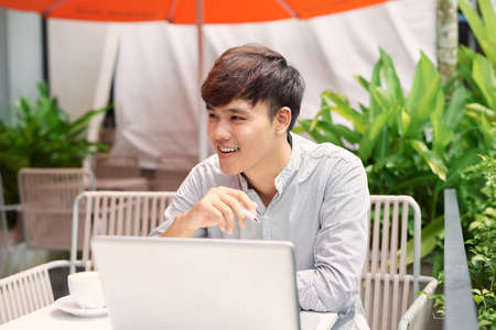 Using advantages of free Wi-Fi. Handsome young man working on laptop and smiling while sitting in sidewalk cafeの写真素材