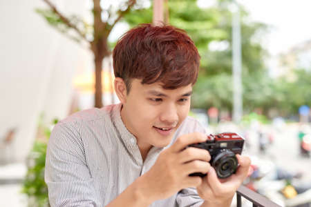 Young serious male sitting at wooden table with photo camera and preparing for photo session while having coffee breakの写真素材