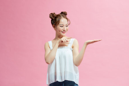 Lady holding palm up and pointing to her right while looking at you smiling in pink backgroundの写真素材