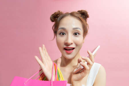 A portrait of young asian woman with colourful shopping bags enjoy mobile technology isolated on pink background.の写真素材